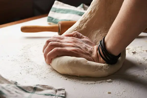 Hands kneading a ball of no yeast pizza dough on a floured surface, with a wooden rolling pin behind