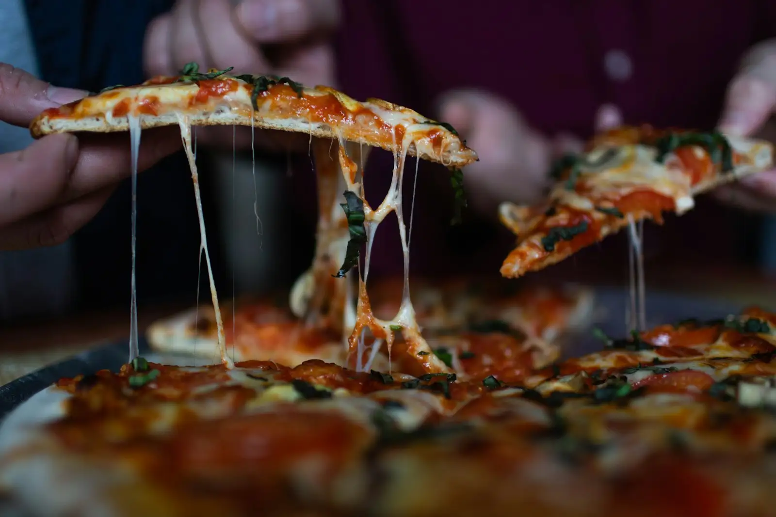 Hands lifting hot thin-crust pizza slices with a gooey cheese pull from a sliced pie