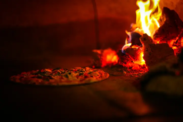 Pizza baking beside live wood flames inside a wood-fired pizza oven