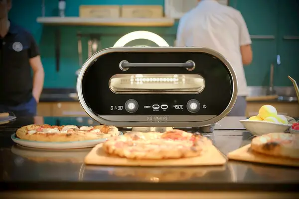 Two finished pizzas on a kitchen counter beside a countertop electric pizza oven