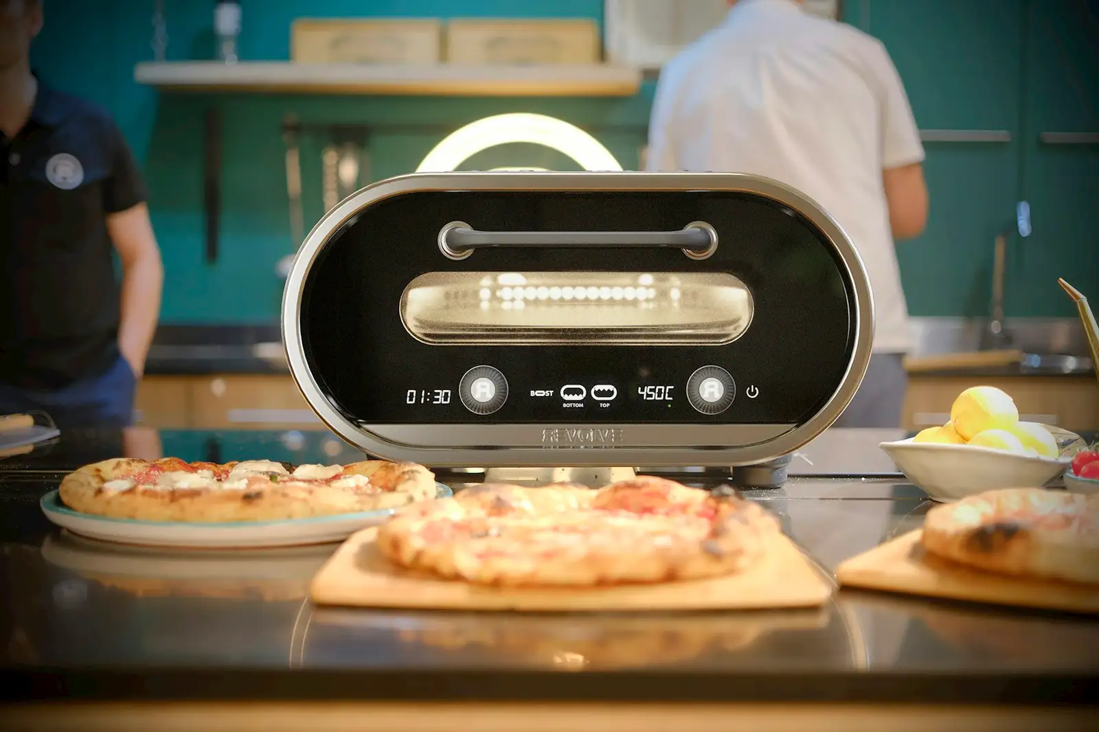 Two finished pizzas on a kitchen counter beside a countertop electric pizza oven