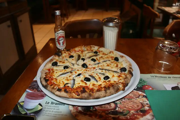 A whole anchovy pizza topped with a radial pattern of anchovy fillets and black olives on a white plate