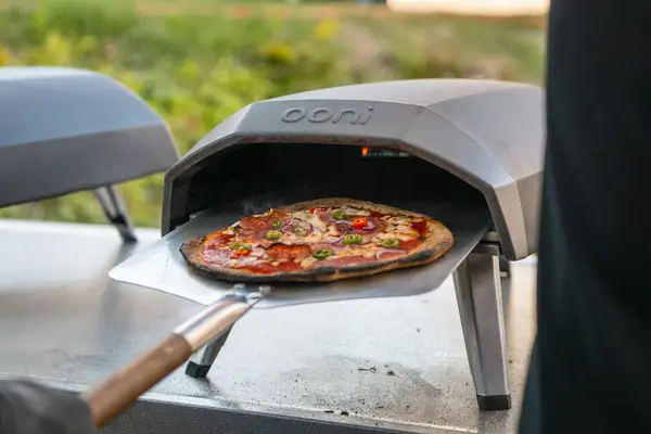 A pizza being launched on a peel into a portable Ooni gas pizza oven in a backyard